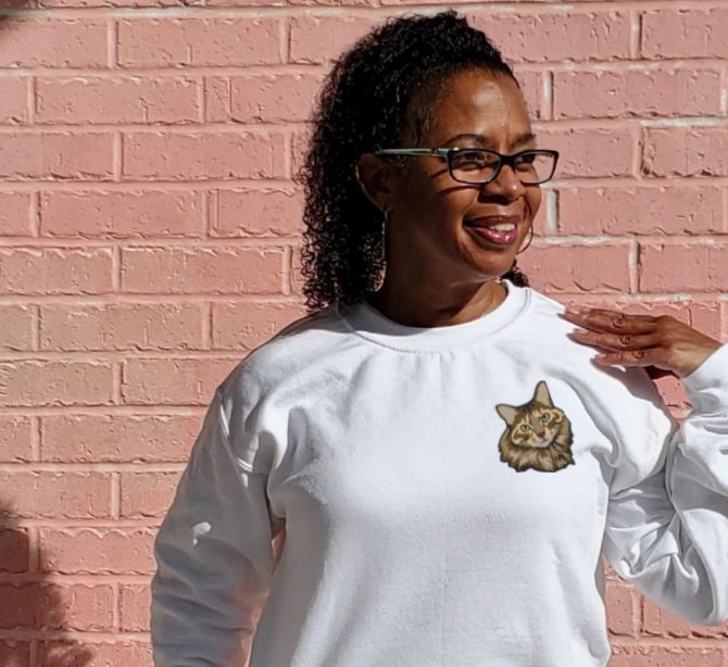 Woman wearing a white crewneck with a brown and black longhaired tabby cat embroidered on the left chest.